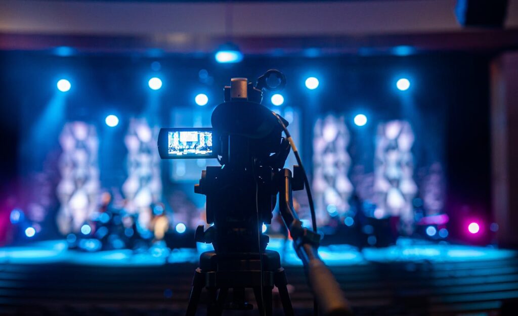 pexels-photo-4889279 Video camera capturing a vibrant concert scene under bright stage lights.